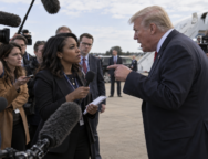 Female journalist faces an aggressive male political figure during an outdoor press gaggle as other reporters look on.