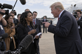 Female journalist faces an aggressive male political figure during an outdoor press gaggle as other reporters look on.